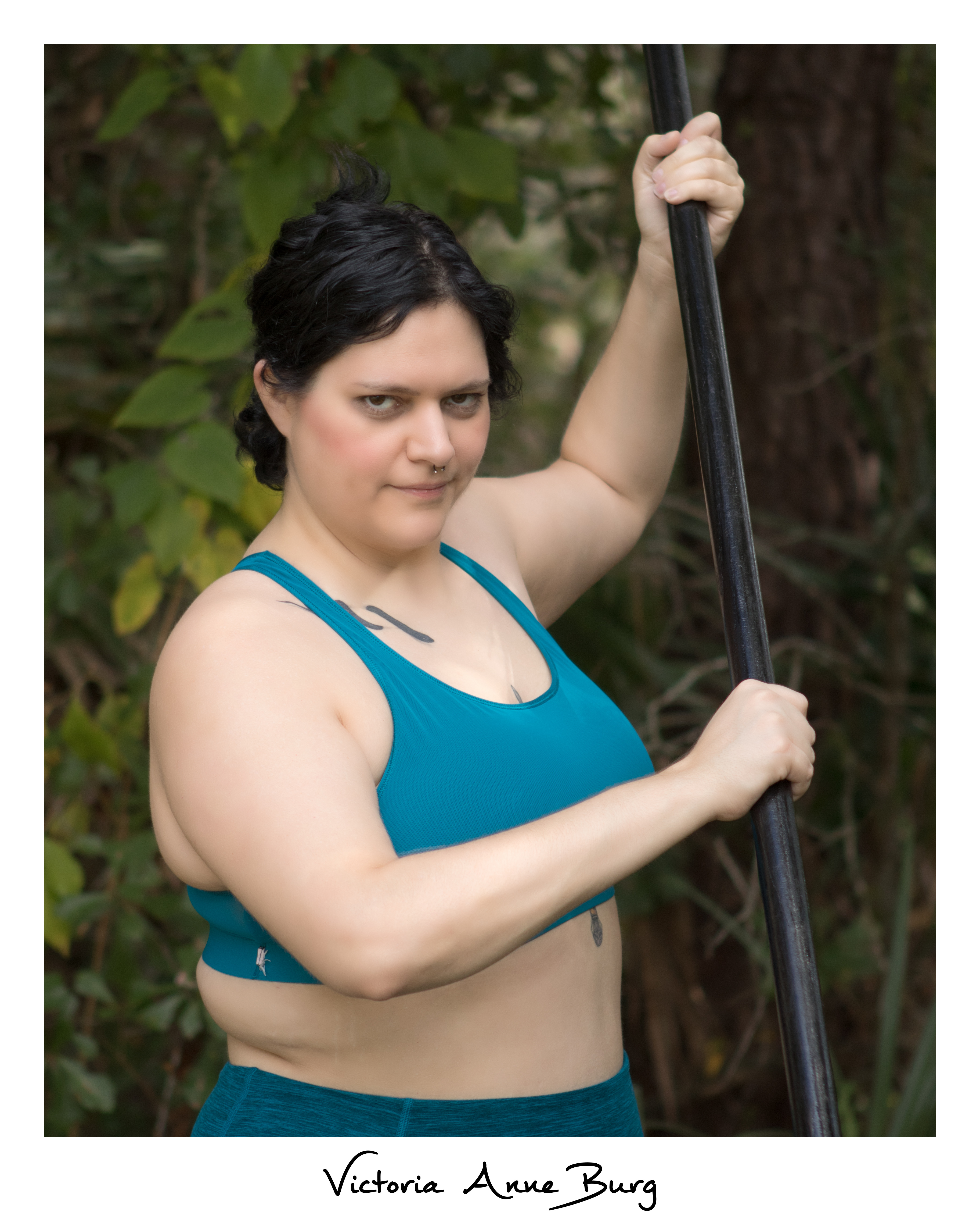Bodyshot of V. Anne Burg, wait-up of black haired person, hair tied back, snake collar bone tattoo, sternum tattoo, holding a black quarter staff, and wearing a turquoise sports bra and pants
