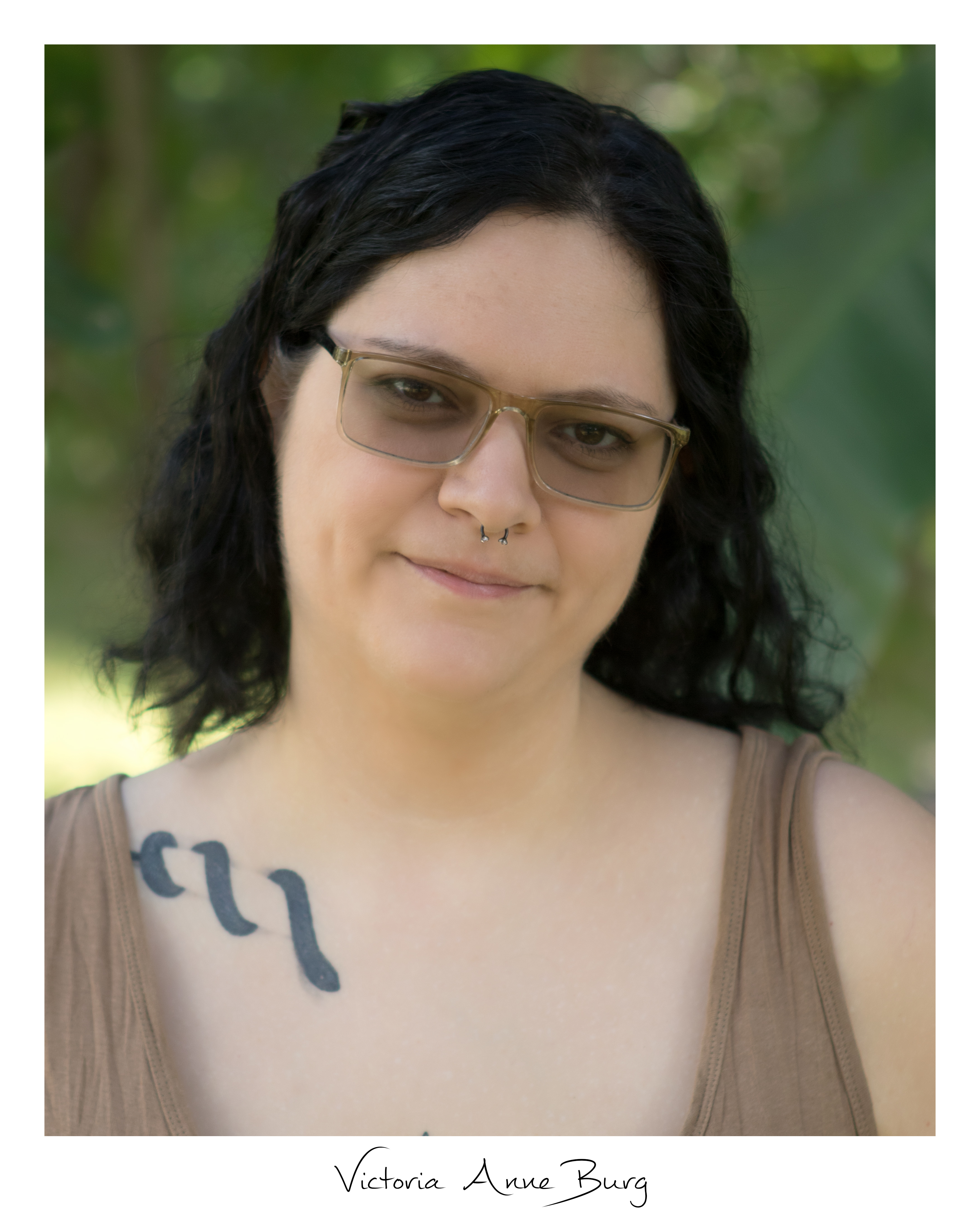 Headshot of V. Anne Burg, closeup of black haired person with glasses, snake collar bone tattoo and wearing tan tank top